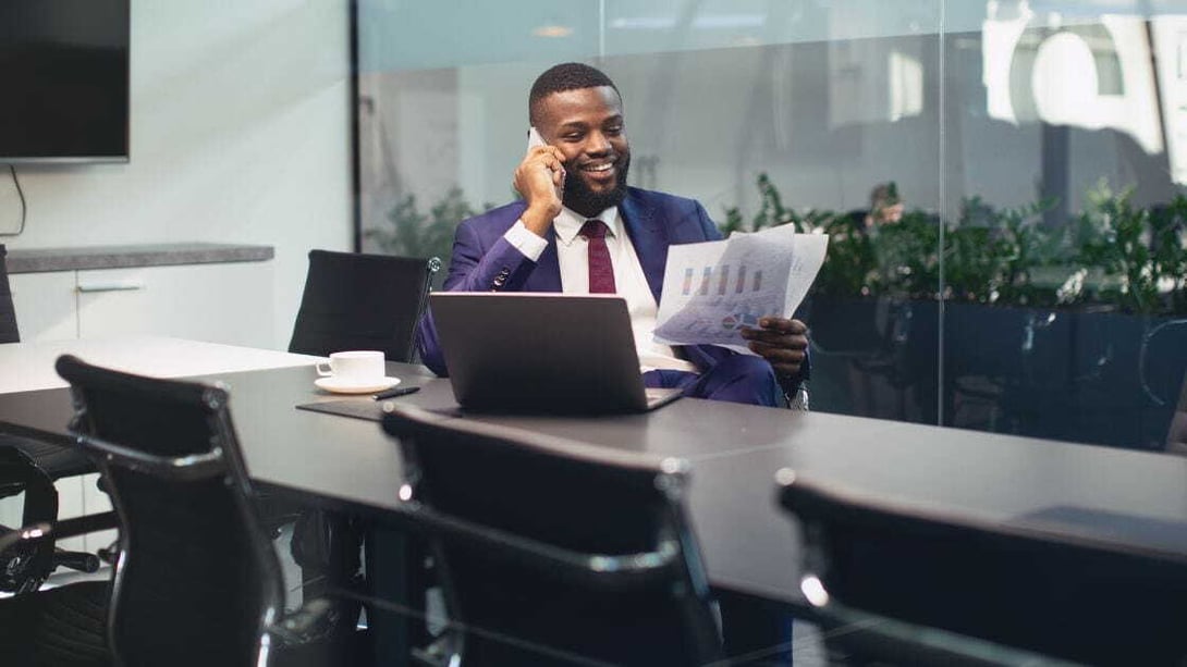 A businessman in an office, talking on the phone and holding printed graphics in his other hand.