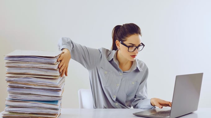 Mulher no escritório e na mesa, muitos papéis à esquerda e um laptop à direita, representando a transformação digital.