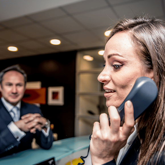 Hotel receptionist answering a customer by IP phone.  There is a customer waiting to be attended to at reception.